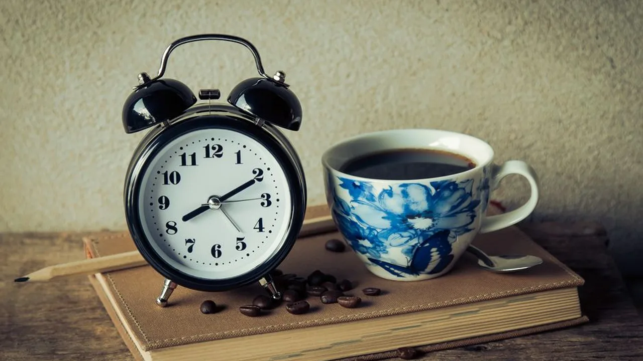 木桌上的筆記本上放著一只黑色雙鈴鬧鐘與一杯裝在青花瓷杯裡的黑咖啡，旁邊散落幾顆咖啡豆