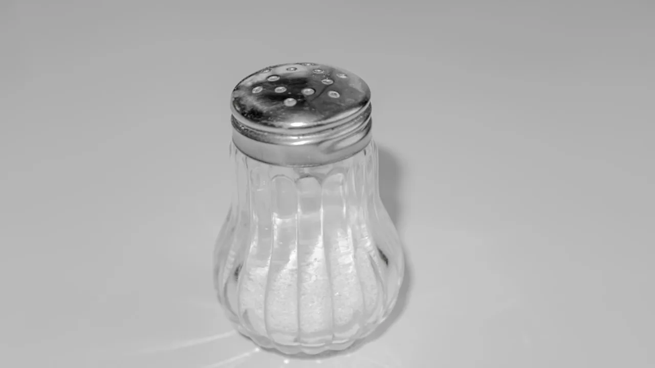 白色檯面上一只金屬蓋的玻璃鹽罐，罐底裝著少量白色鹽粒（黑白照片）